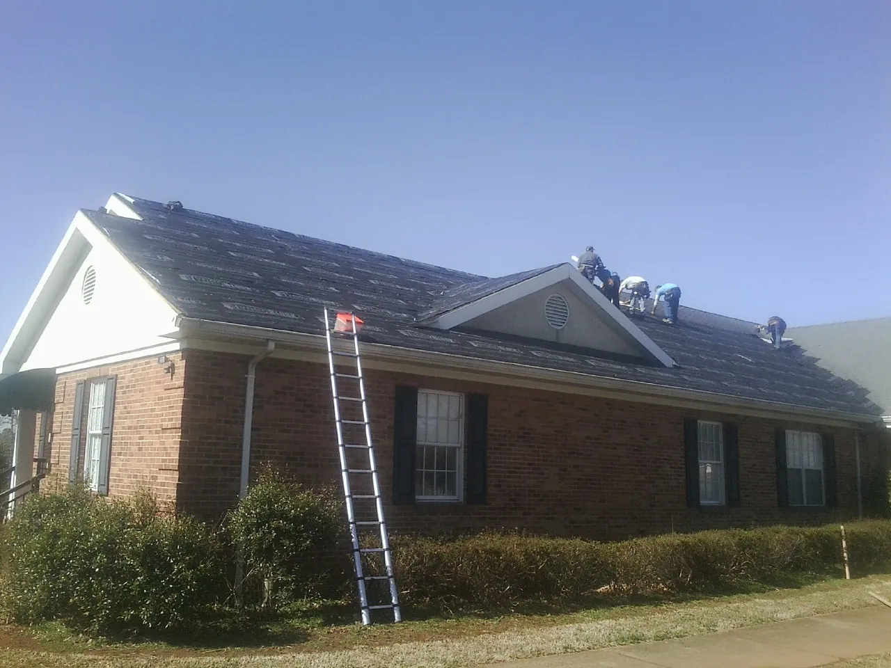 Crew tearing shingles off a brick ranch roof with a ladder set — Emergency Roof Repair work by our crew in Armstrong Creek
