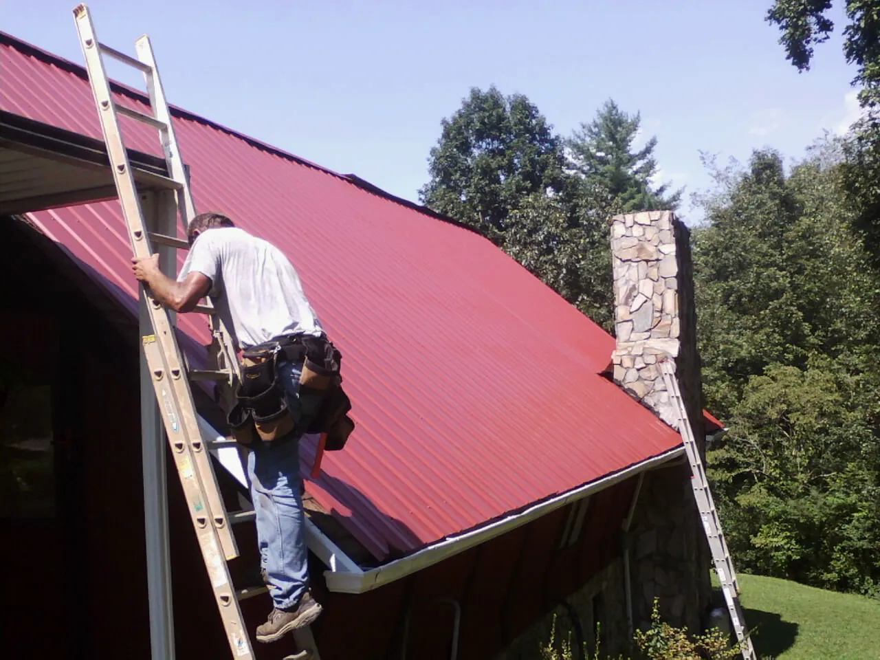 Roofer on a ladder beside a red metal roof and stone chimney — Soffit & Fascia Repair by Roofing Brothers in Armstrong Creek